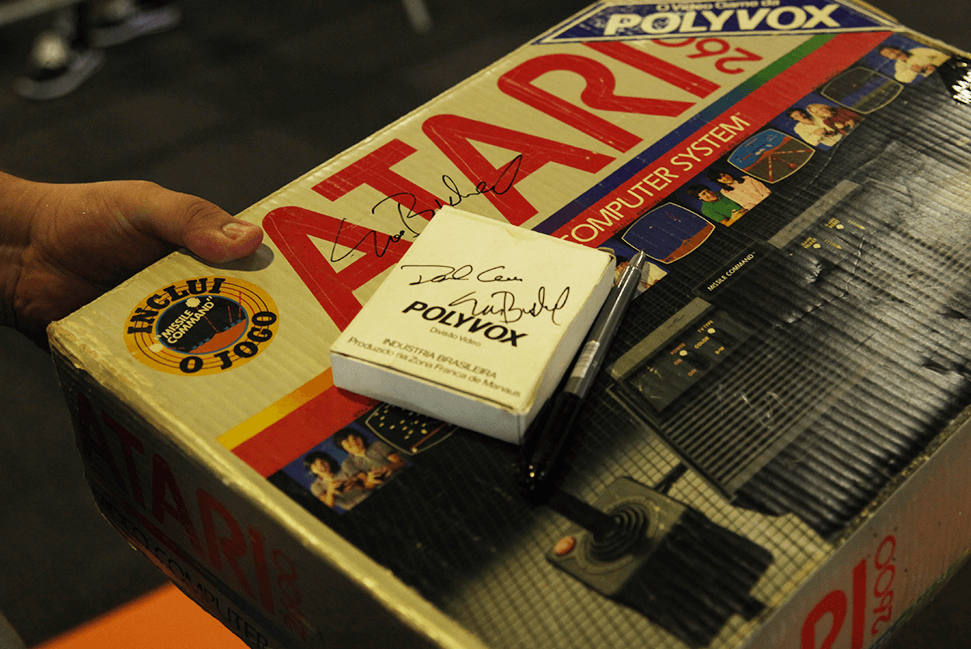 Caixa de um video game Atari, autografada por personalidades importantes pela criação do mesmo. Em cima da caixa tem uma caneta e outra caixa menor de um Polybox, também autografada.