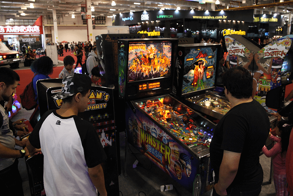 Fotografia de uma máquina de fliperama do Pacman, ao lado duas duas máquinas de pinball. Todas com pessoas jogando.