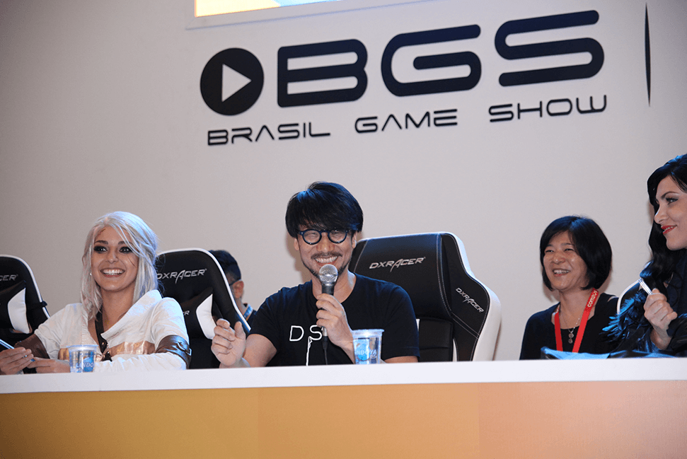 Fotografia do desenvolvedor de Metal Gear, Kojima, sorrindo e segurando um microfone. Ele está sentado no painel da BGS.